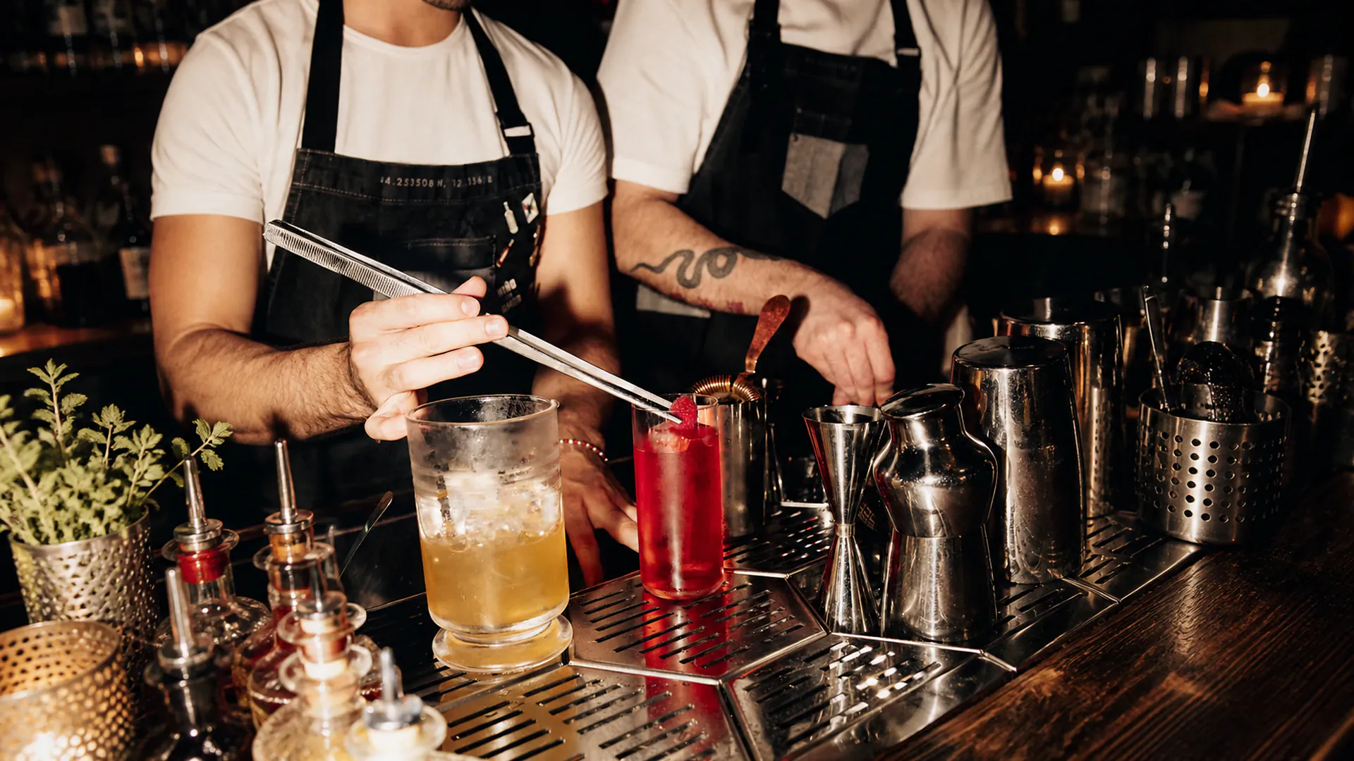 Deux barmen en tablier noir dressent deux cocktails sur le comptoir, entourés de shakers, de doseurs et de flacons à bec verseur.