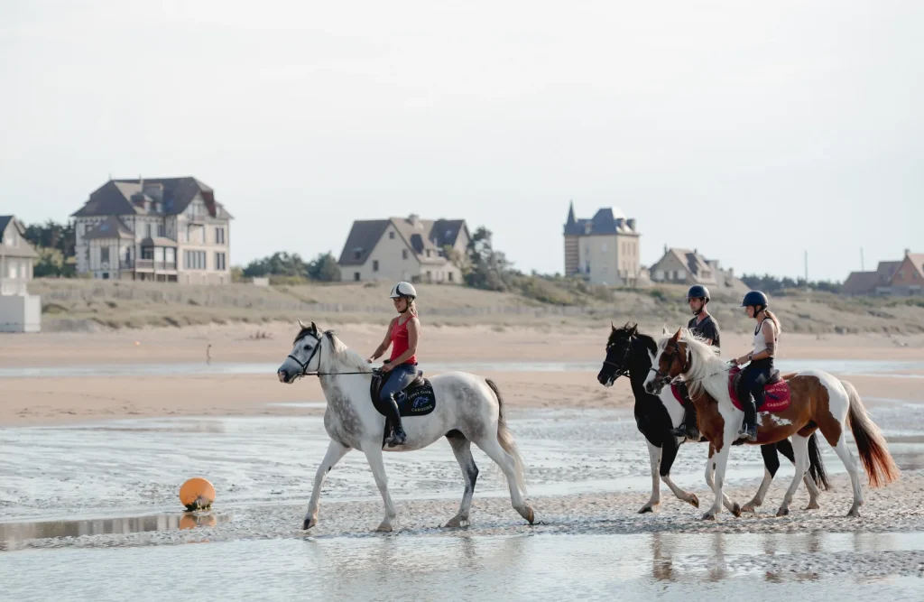 Quatre cavaliers casqués avancent au pas sur le sable mouillé d'une plage de la Côte Fleurie, devant les villas normandes du front de mer.