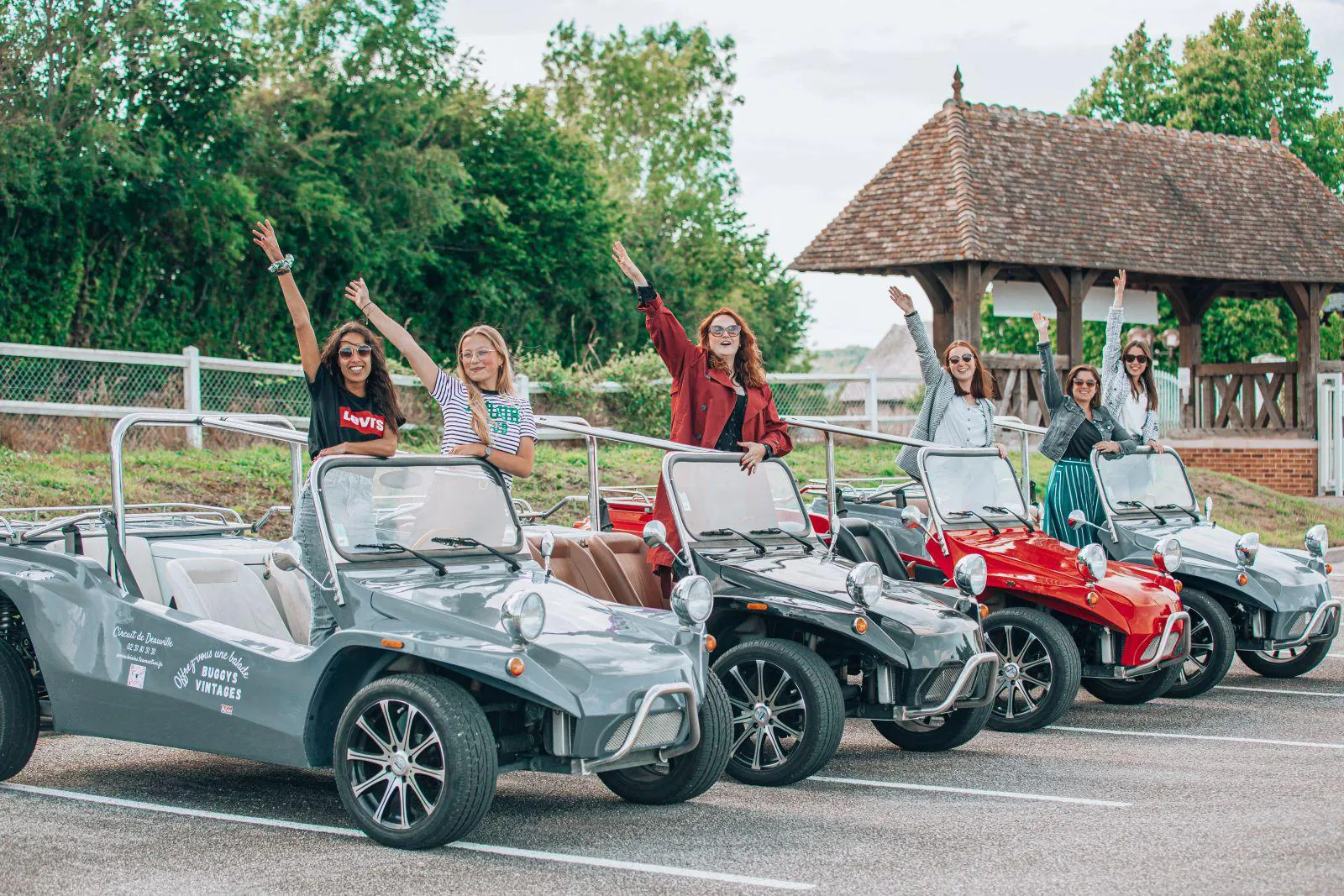 Six participantes debout dans quatre buggys vintage alignés, bras levés, devant un lavoir normand à toit de tuiles.