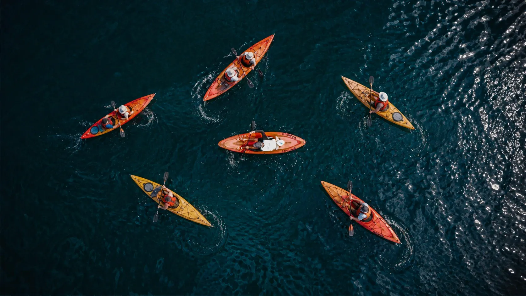 Vue aérienne d'une eau bleu sombre : trois nageurs en crawl laissent un long sillage d'écume, escortés à distance par un kayak orange et son pagayeur.