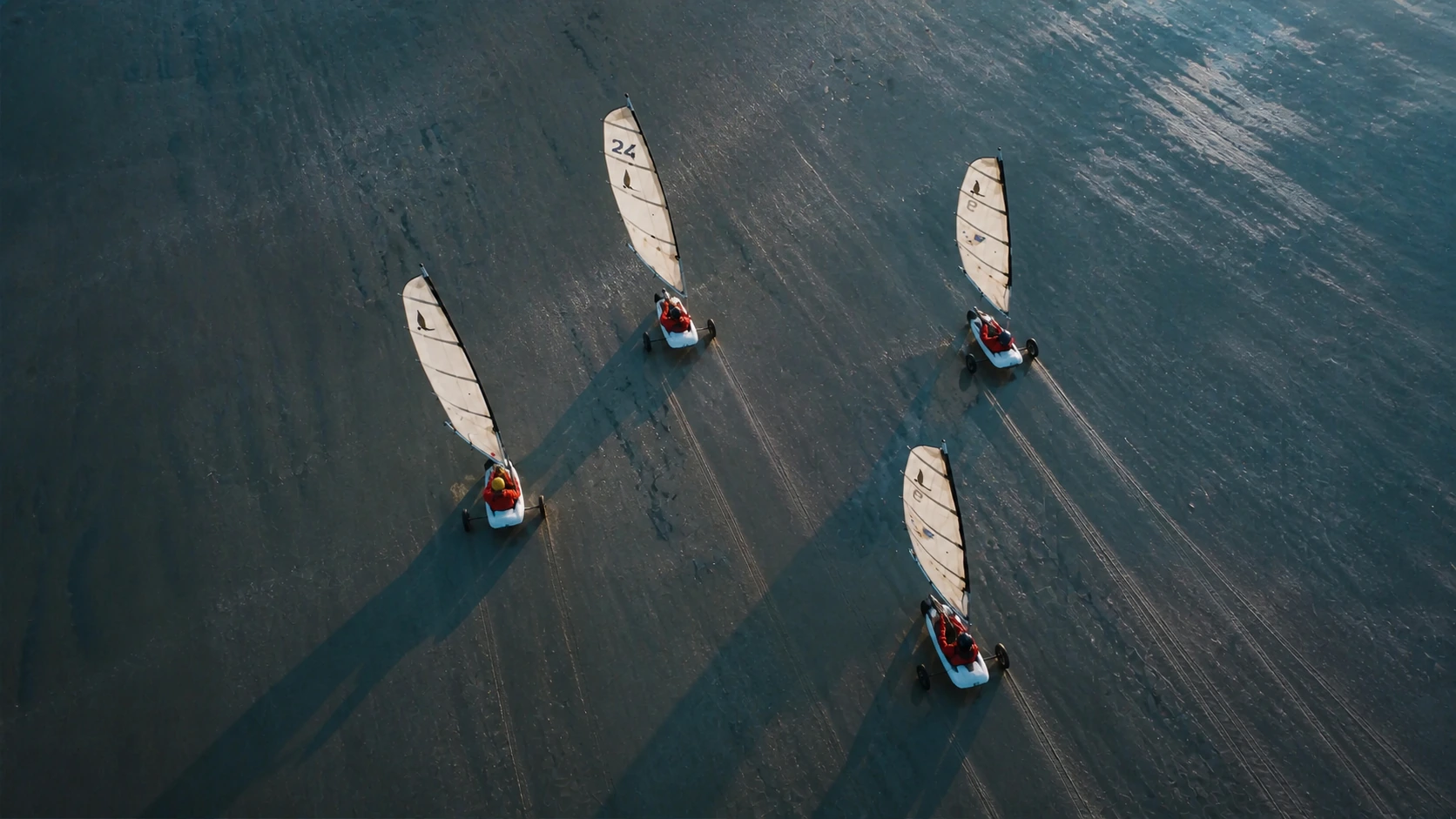 Quatre chars à voile en ligne sur le sable humide d'une grande plage, vus du ciel, leurs ombres allongées par le soleil bas.
