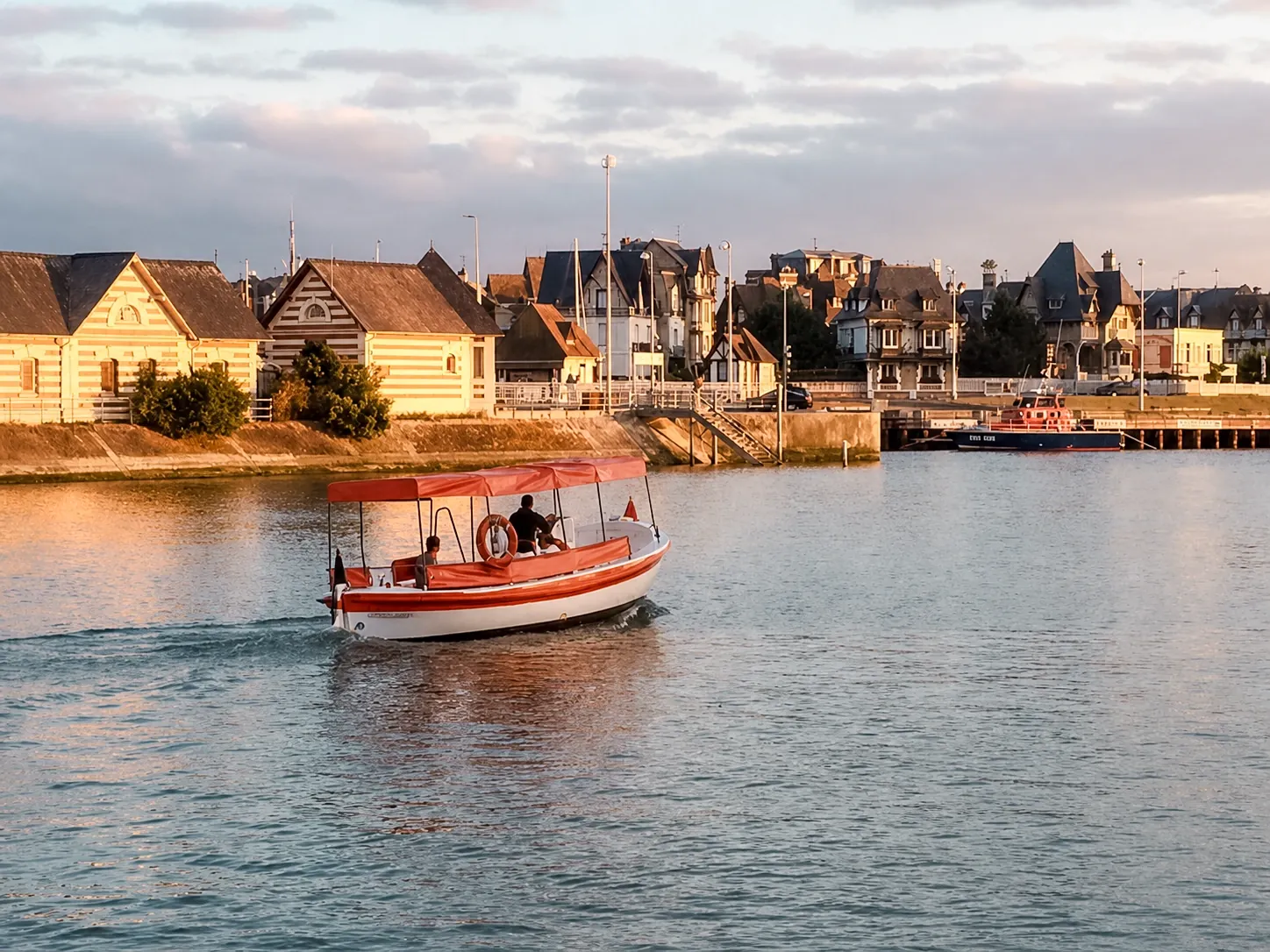Le bac de Deauville-Trouville traversant la Touques au soleil couchant, sa bâche rouge tendue au-dessus des passagers, les cabines de bois et les villas normandes de la rive en arrière-plan.