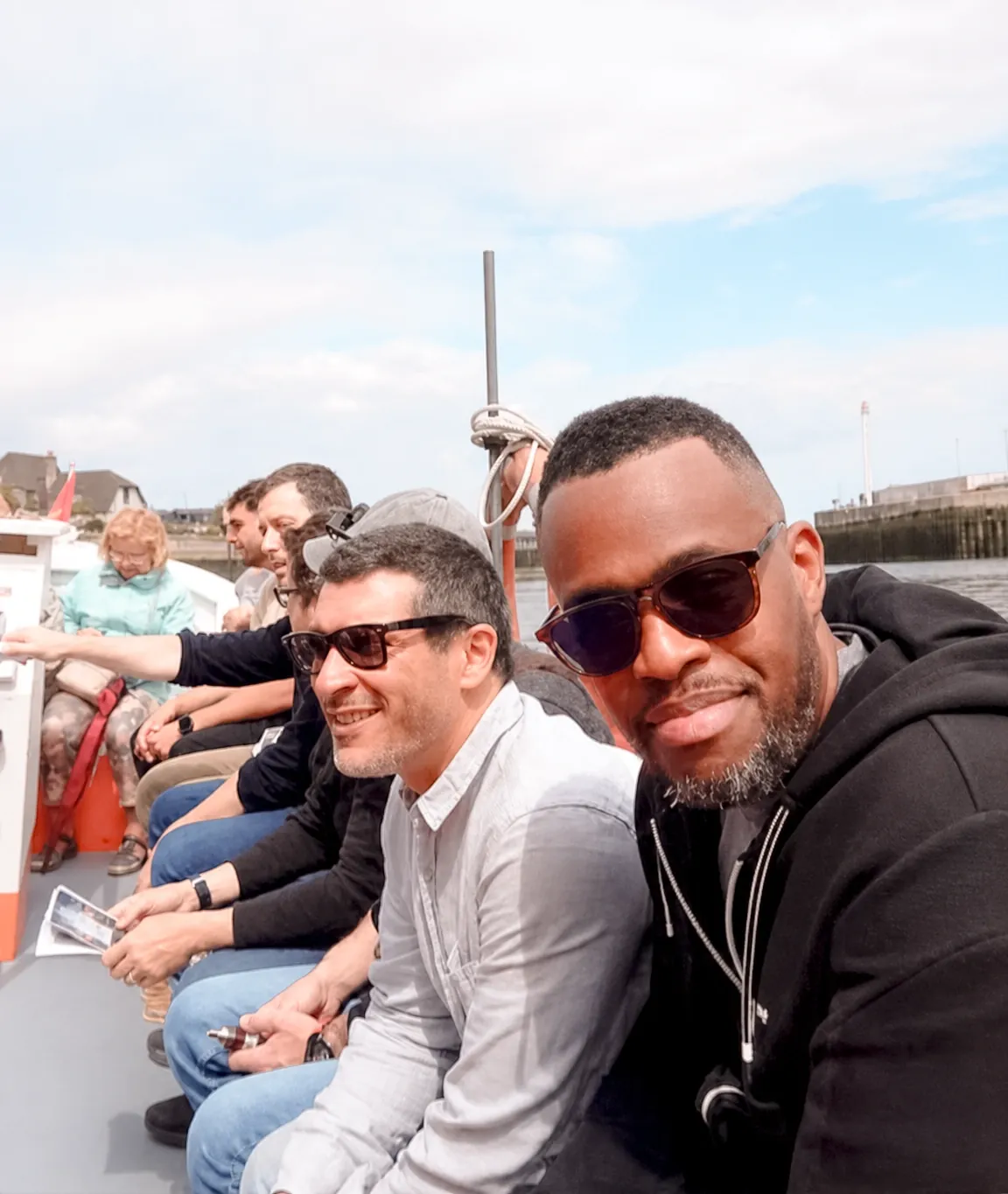 Des participants assis en rangée sur le banc du bac pendant la traversée, lunettes de soleil et feuille de route à la main, la jetée au loin.