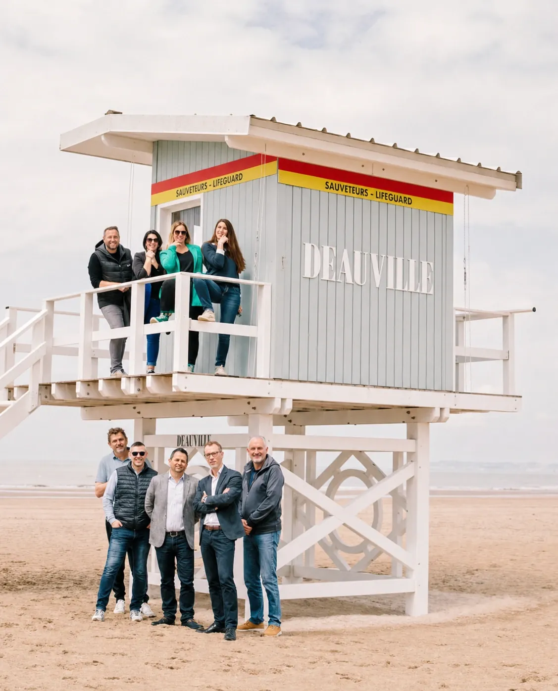 Une équipe répartie autour d'un poste de sauveteurs de la plage de Deauville, quatre personnes sur la passerelle et cinq au pied des pilotis, le nom de la ville peint sur le bardage.