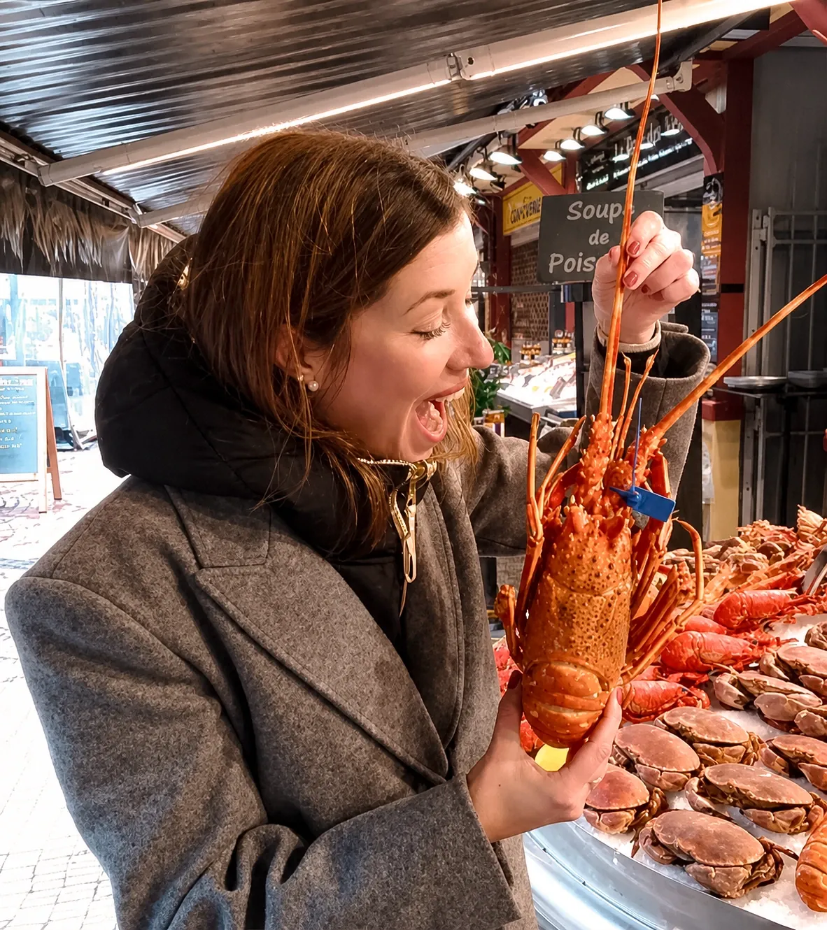 Une participante brandit en riant un homard cuit devant l'étal d'un poissonnier du marché, tourteaux et crustacés alignés sur la glace.