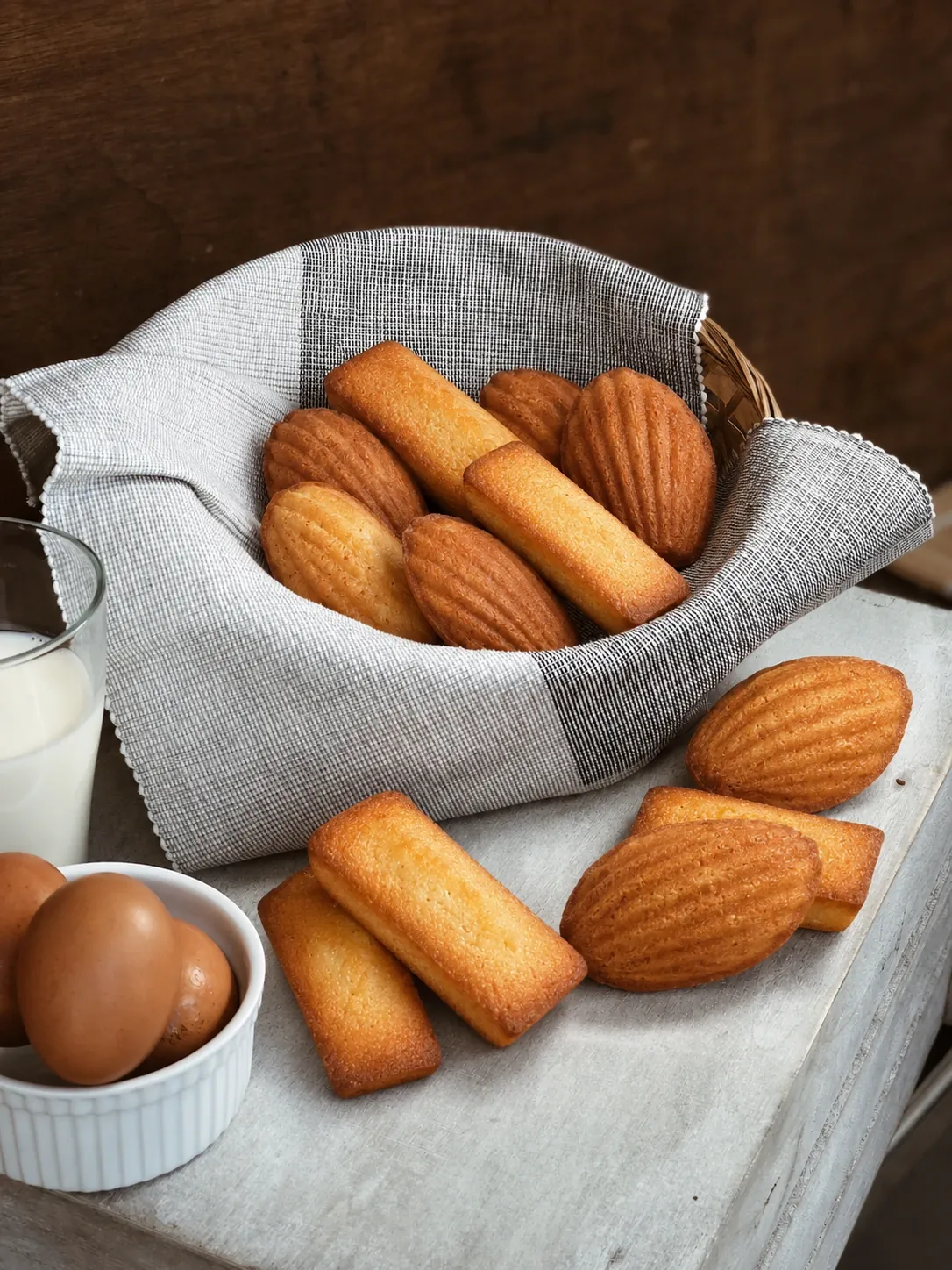 Un panier de madeleines et de financiers doublé d'un torchon, posé sur une planche de bois avec un verre de lait et des œufs frais.