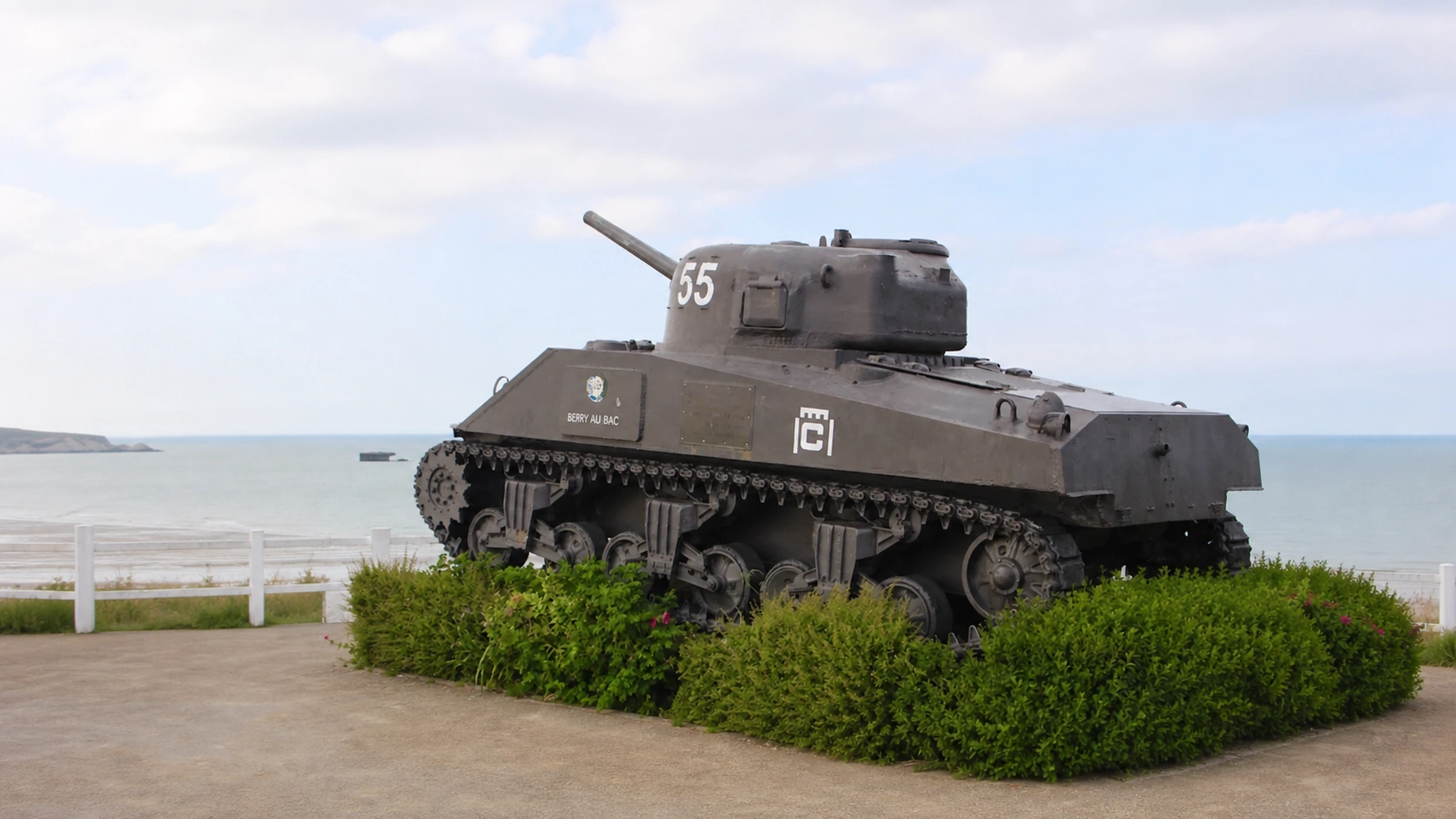 Un char Sherman conservé face à la mer sur le front de mer normand, un caisson du port artificiel du Débarquement visible au large.