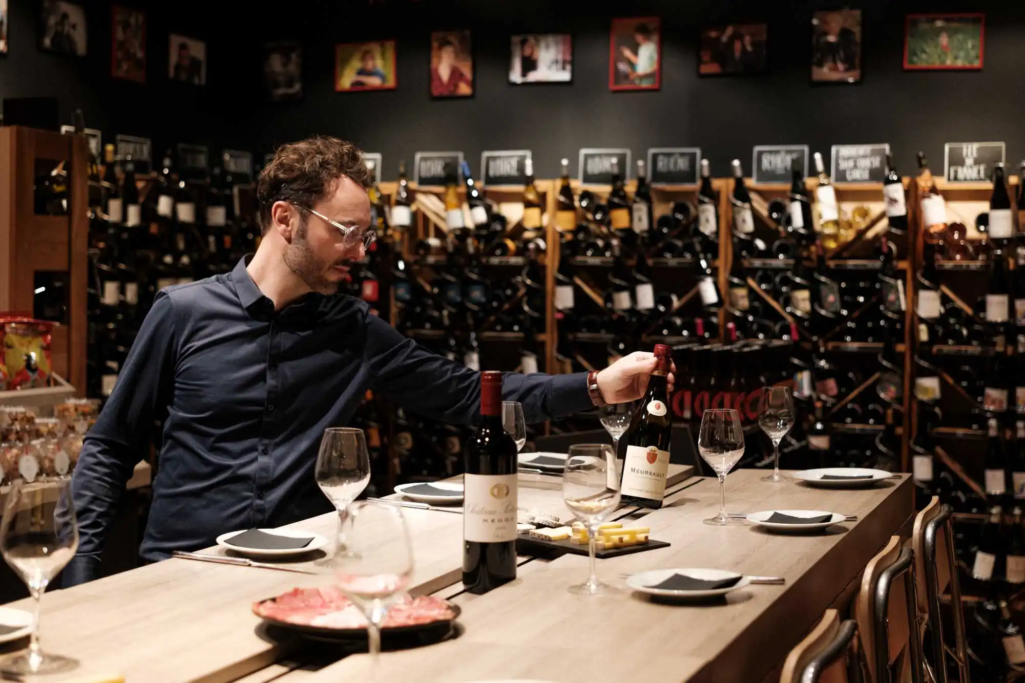 Dans une cave, un caviste dispose une bouteille sur une longue table dressée de verres et d'assiettes, devant des rayonnages de vins.