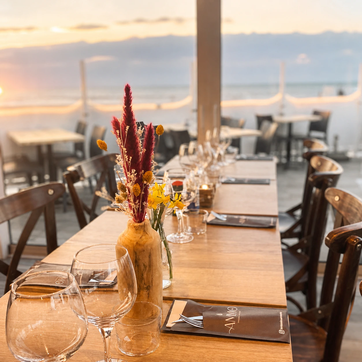 Une grande tablée dressée sur la terrasse d'un restaurant de bord de mer au crépuscule : plateaux de bois, verres à pied, bouquet de fleurs séchées et menus posés à chaque place.
