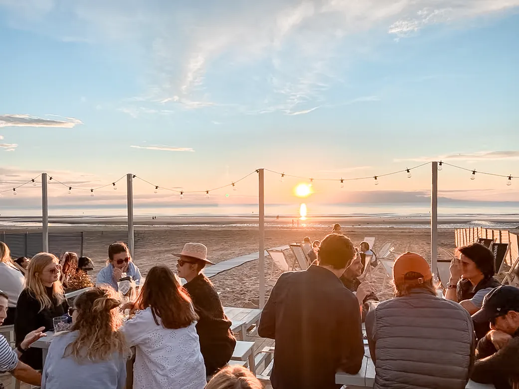 Dîner sur la plage au coucher du soleil : convives attablés sous des guirlandes lumineuses, transats et sable face à la mer.