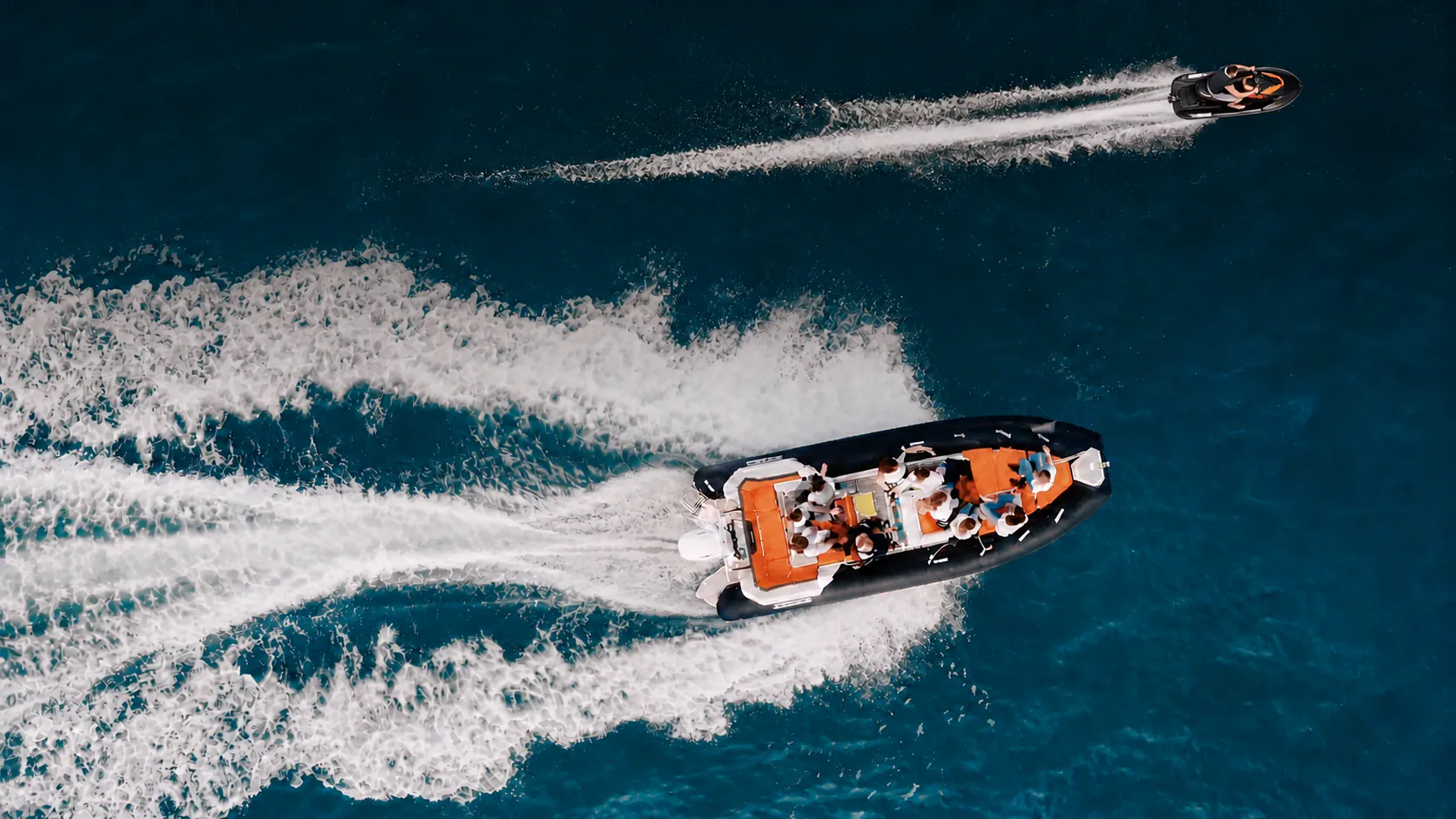 Vue aérienne d'un zodiac chargé de participants et d'un jet-ski lancés à pleine vitesse, laissant deux longs sillages d'écume.