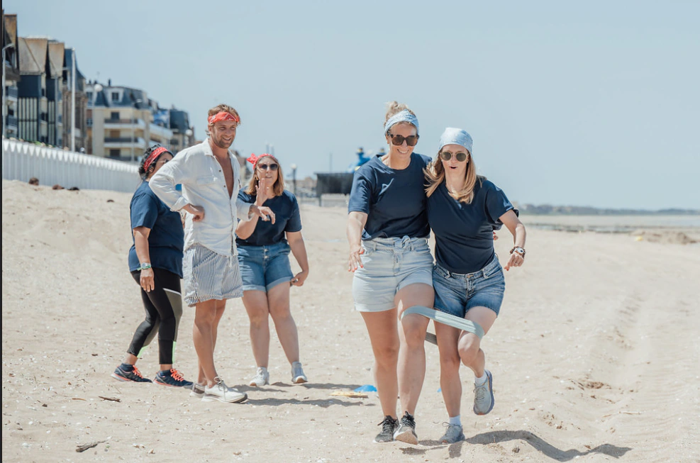 Épreuve de course à deux, jambes attachées, sur la plage de Deauville : deux participantes en T-shirt marine progressent dans le sable, encouragées par le reste de l'équipe, front de mer en arrière-plan.