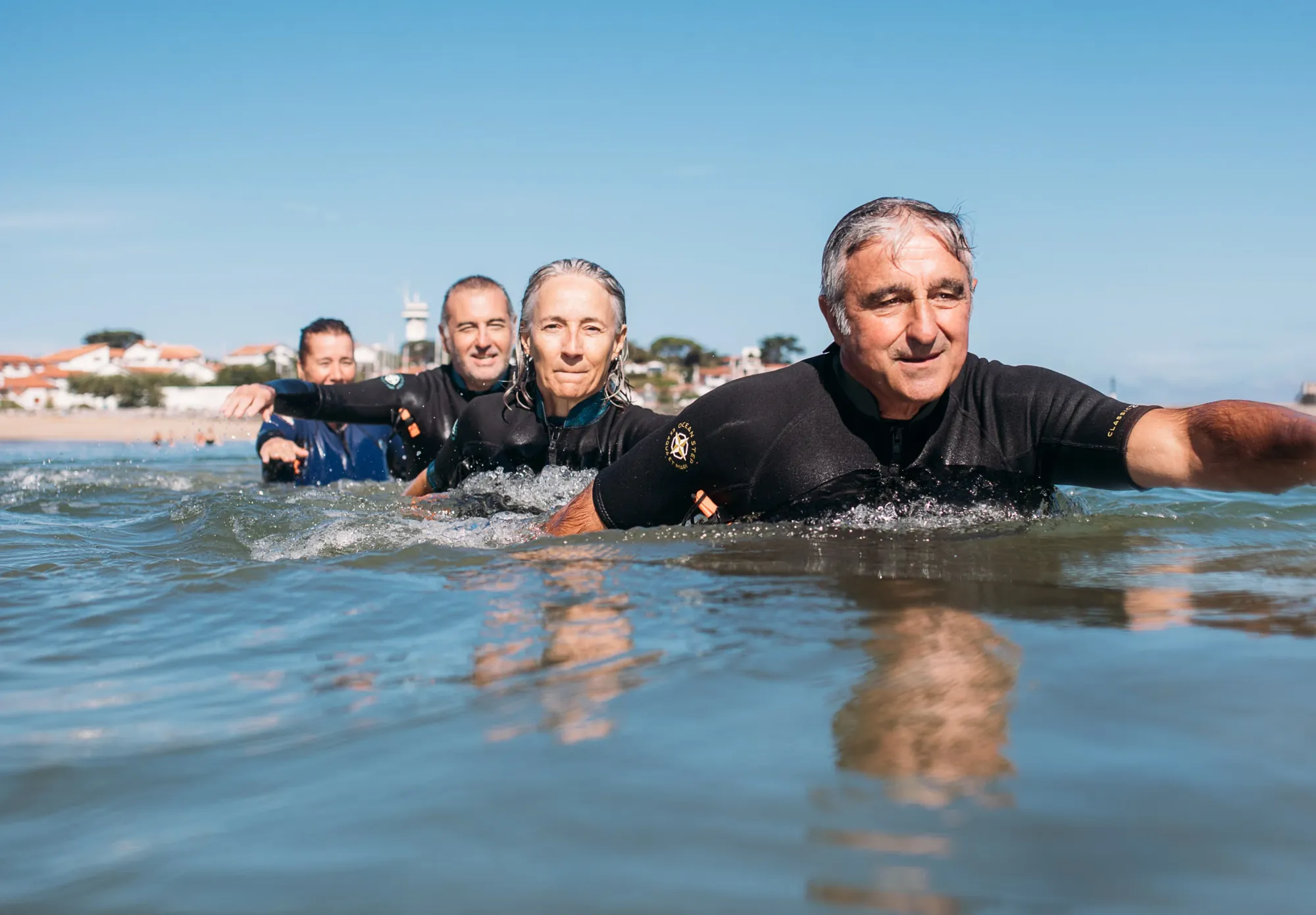 Quatre pratiquants en combinaison néoprène avancent en ligne dans l'eau jusqu'à la taille, bras tendus, le front de mer derrière eux.