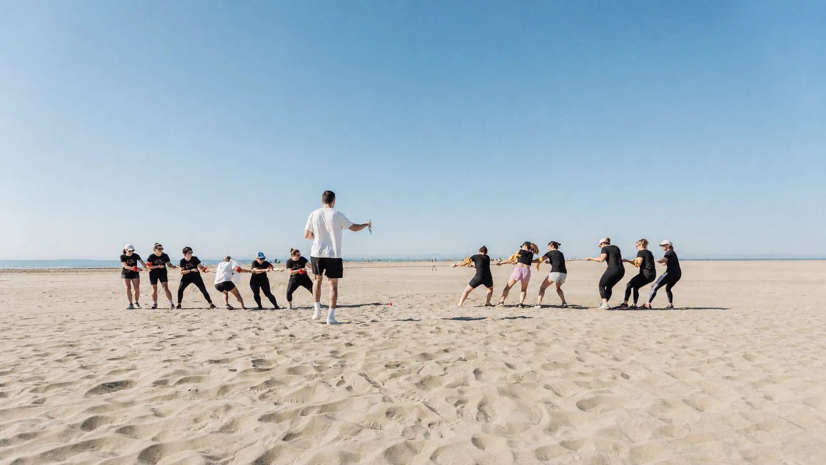 Épreuve de tir à la corde sur la plage : deux équipes tirent de part et d'autre, un arbitre au centre, la mer à l'horizon.