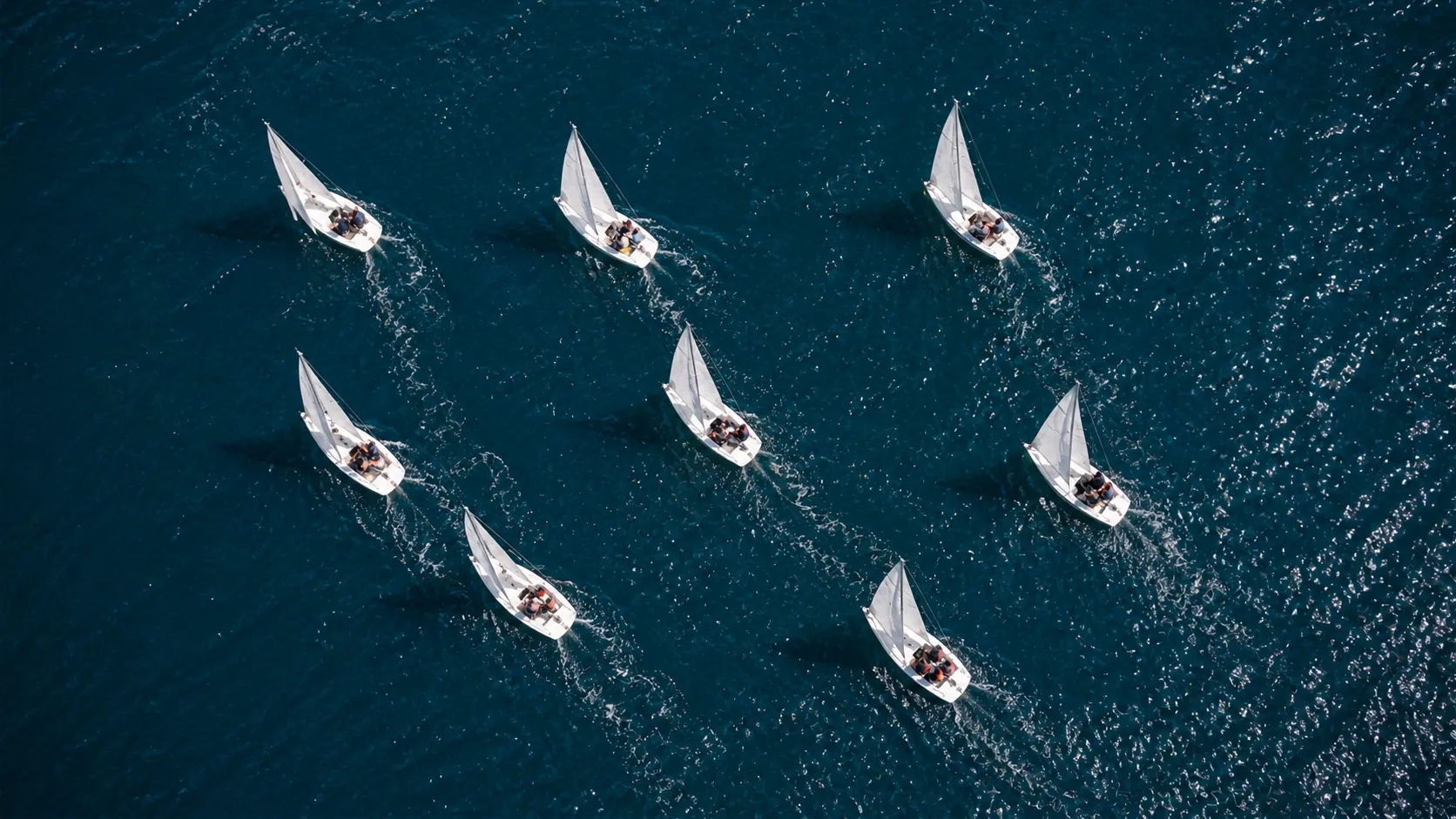 Vue aérienne de huit voiliers blancs en régate sur une mer bleu profond, chacun avec son équipage à bord, laissant derrière lui un sillage d'écume.