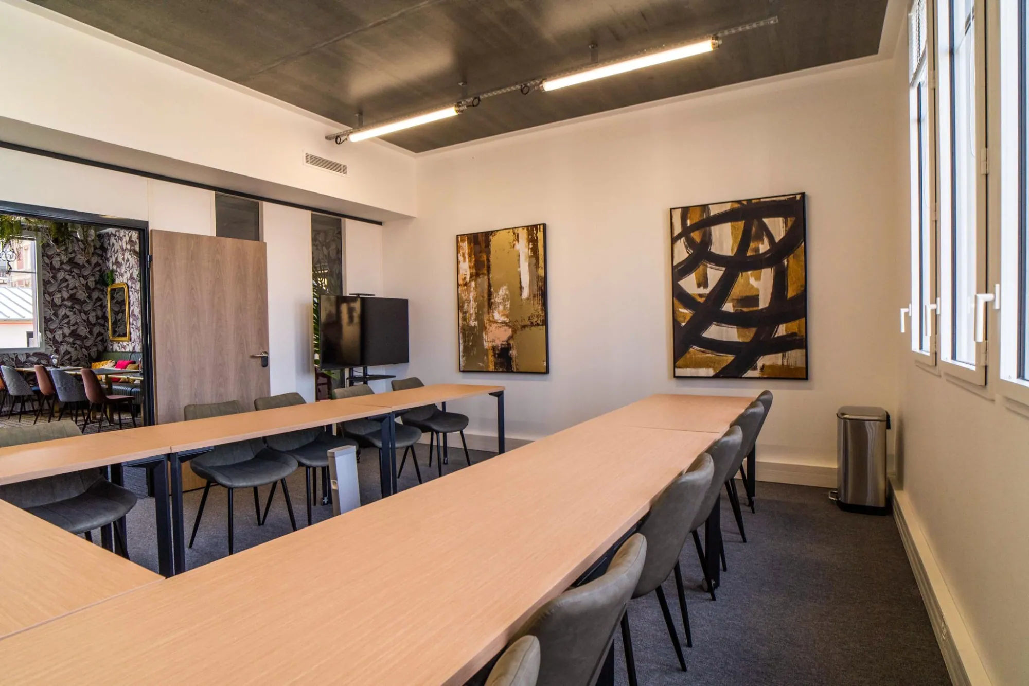Salle de réunion : deux longues tables en bois clair disposées en L, chaises grises, écran mural et deux toiles abstraites, lumière du jour par les fenêtres.