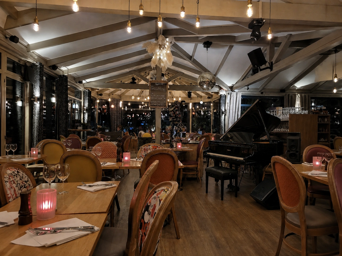 Une salle de restaurant le soir : charpente claire, ampoules à filament suspendues, tables dressées éclairées à la bougie, un piano à queue et une boule à facettes au fond.