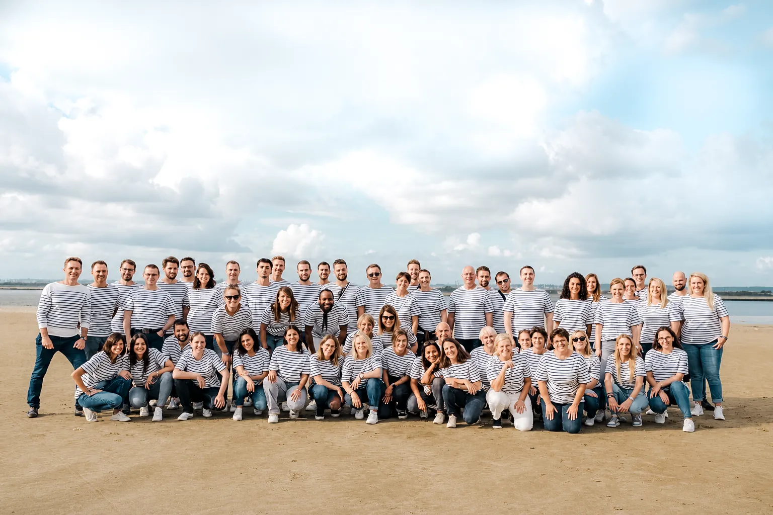 Photographie de groupe sur la plage : une cinquantaine de collaborateurs en marinière rayée, debout et accroupis sur trois rangs, devant la mer et un ciel nuageux.