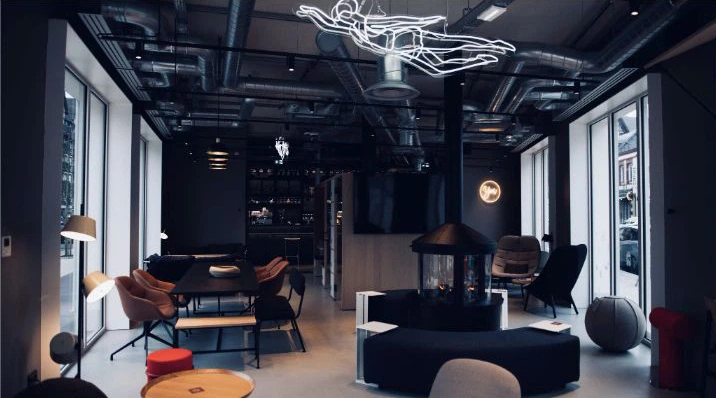 Une salle au style industriel : plafond technique apparent, grandes baies vitrées, cheminée centrale, tables et fauteuils bas répartis autour.