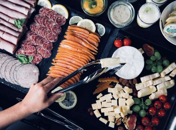 Un grand plateau de brunch vu du dessus : charcuterie roulée, saumon fumé en éventail, camembert, cubes de fromage, tomates cerises et raisin, avec une pince saisissant une part de fromage.