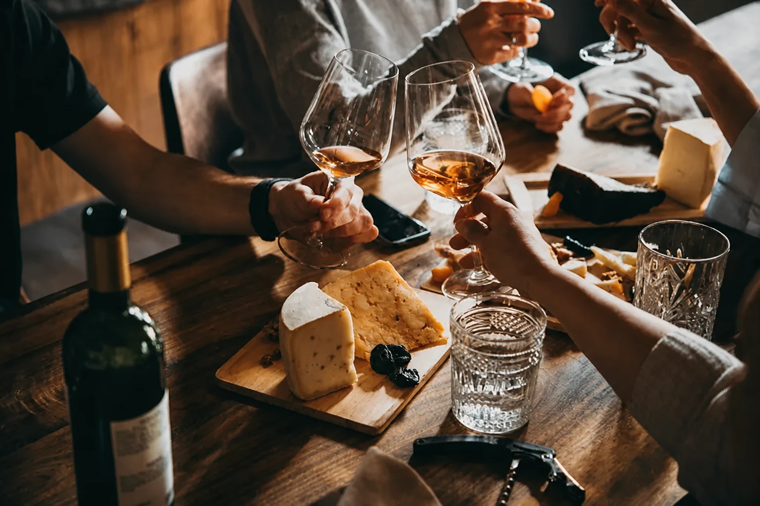 Deux mains trinquent avec des verres de calvados au-dessus d'une planche de fromages, de pruneaux et de noix, une bouteille posée au premier plan.