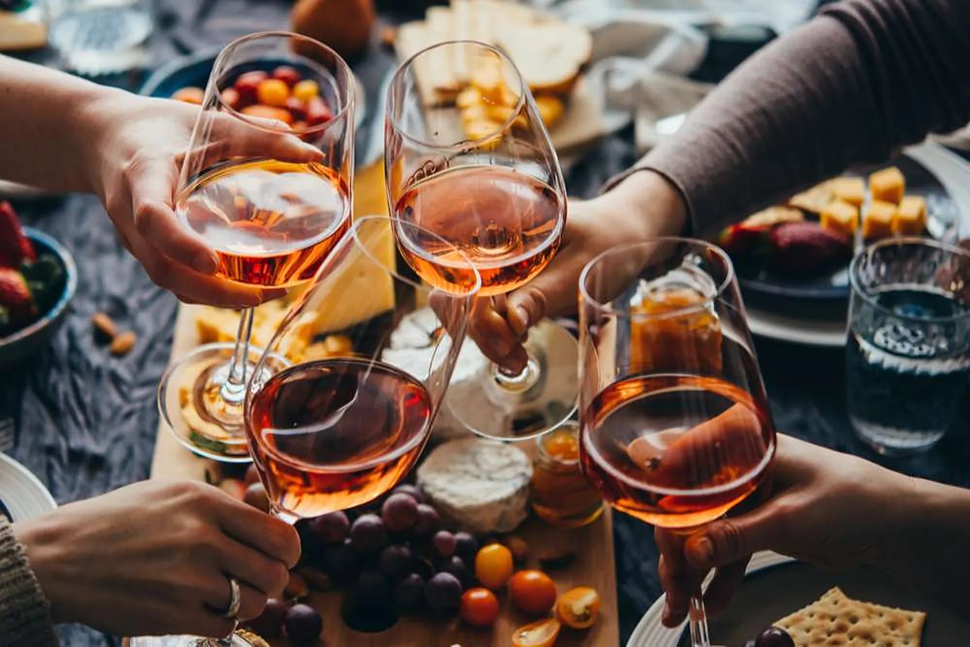 Quatre verres de rosé trinquent au-dessus d'une planche de fromages, de charcuterie et de fruits.