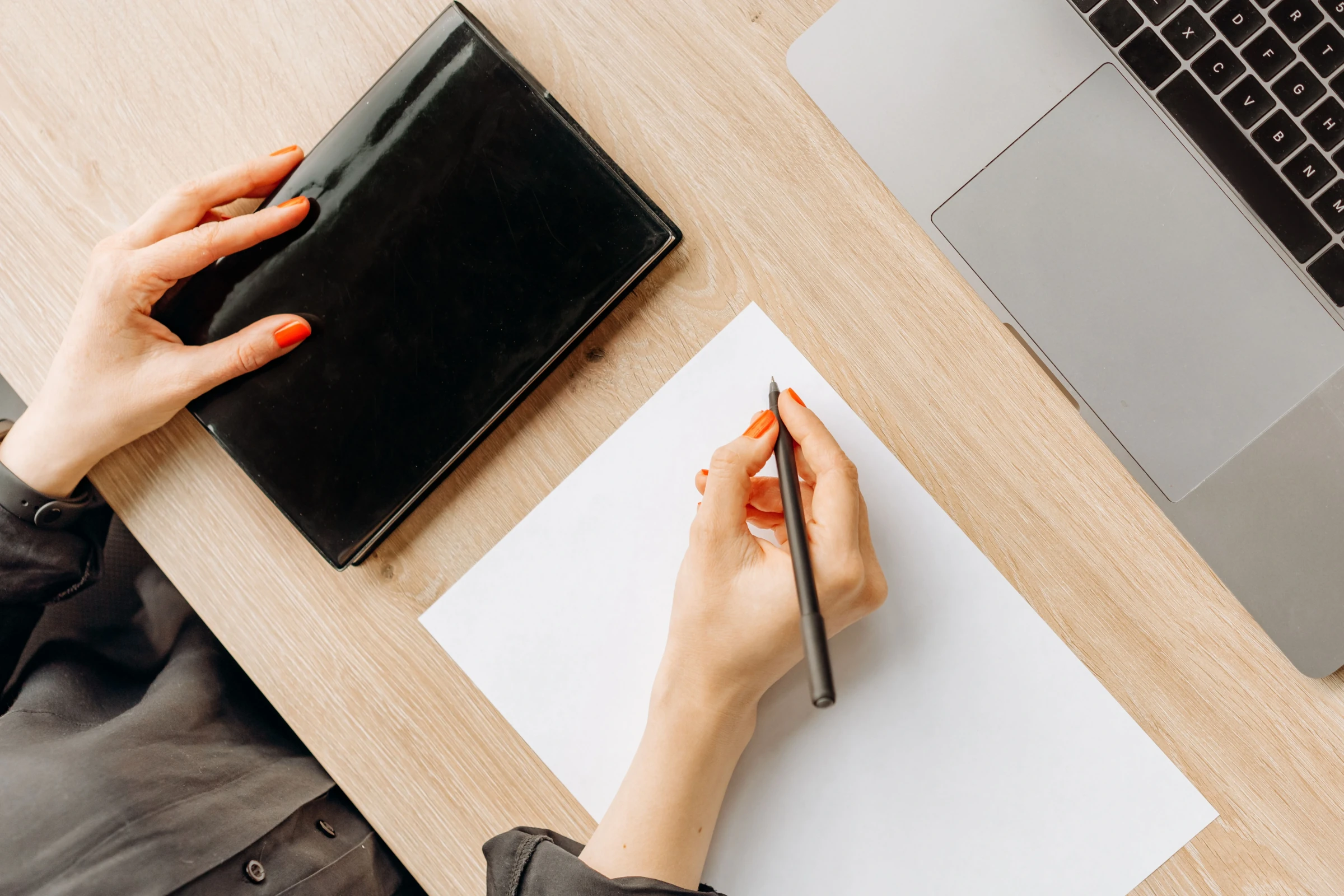 Vue de dessus d'un bureau : une main posée sur un carnet noir fermé, l'autre tenant un stylo au-dessus d'une feuille blanche, à côté d'un ordinateur portable.