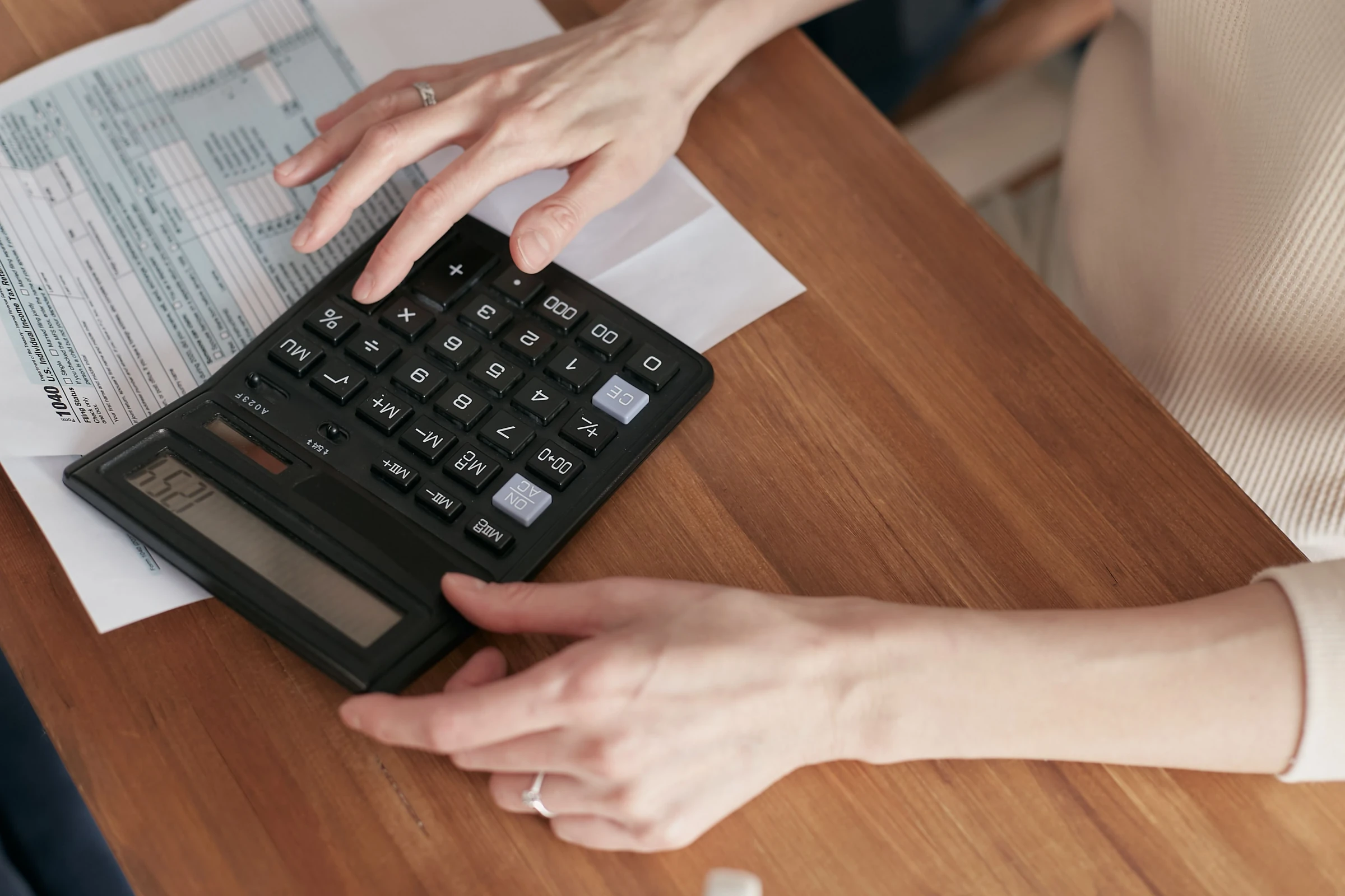 Deux mains sur une calculatrice de bureau posée sur des formulaires imprimés, l'écran affichant un total.