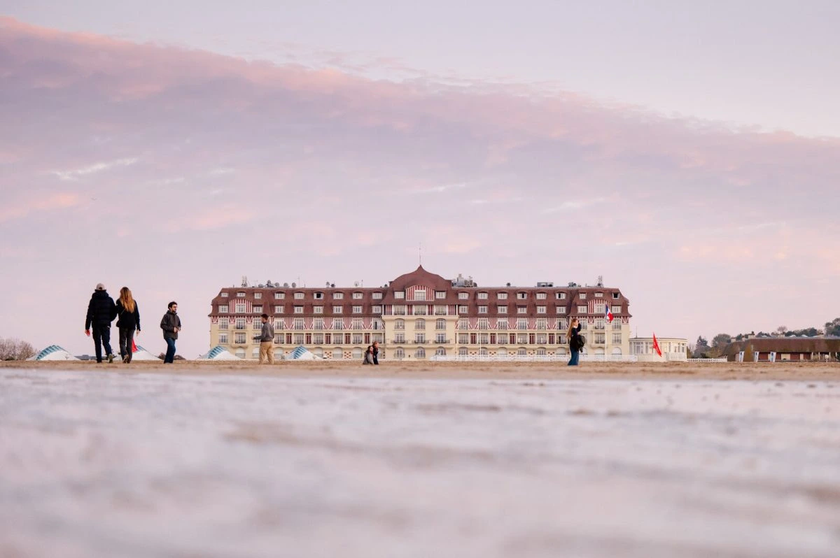 Un grand hôtel de front de mer sous un ciel rose au crépuscule, quelques promeneurs sur le sable devant les tentes de plage repliées.