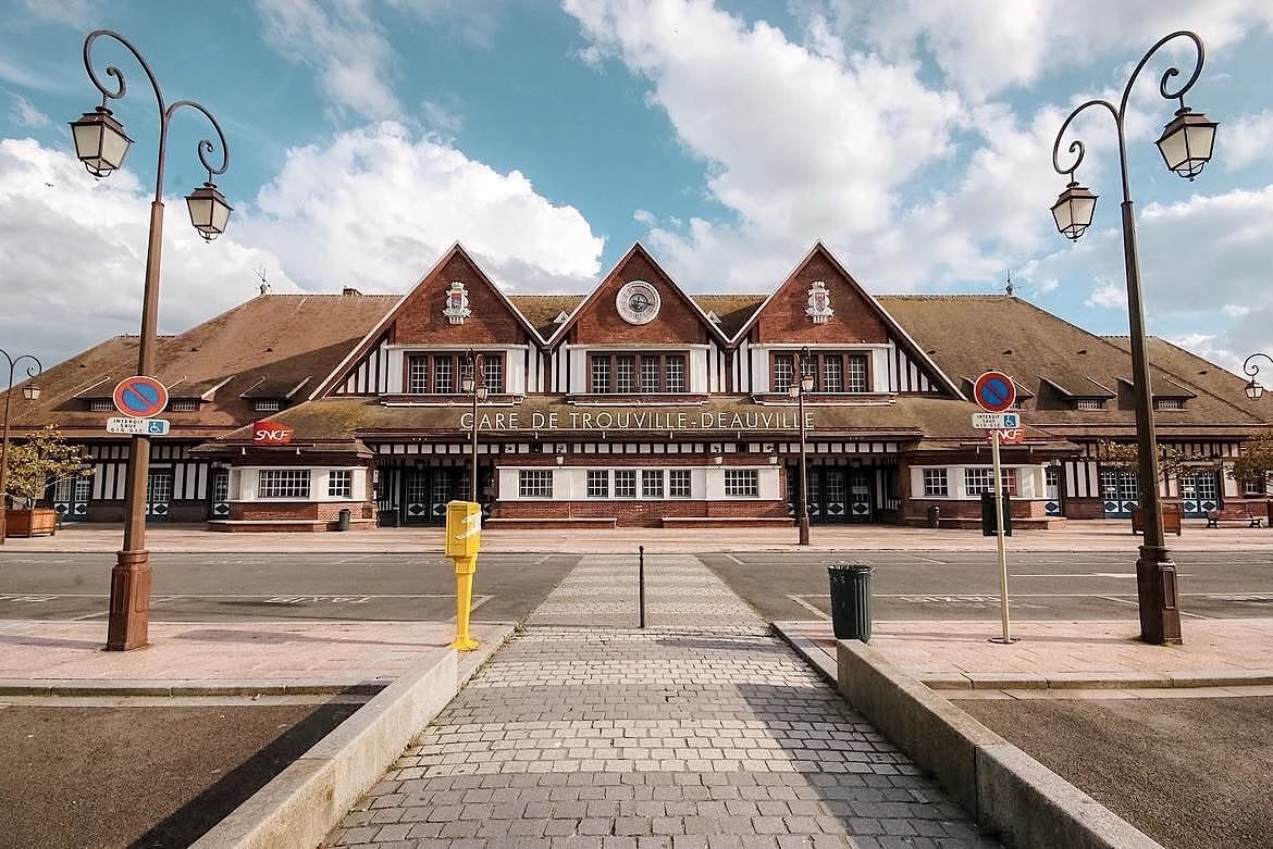 La façade de la gare de Trouville-Deauville, briques rouges et pans de bois sous trois pignons, son horloge et son nom en lettres blanches, devant le parvis désert.