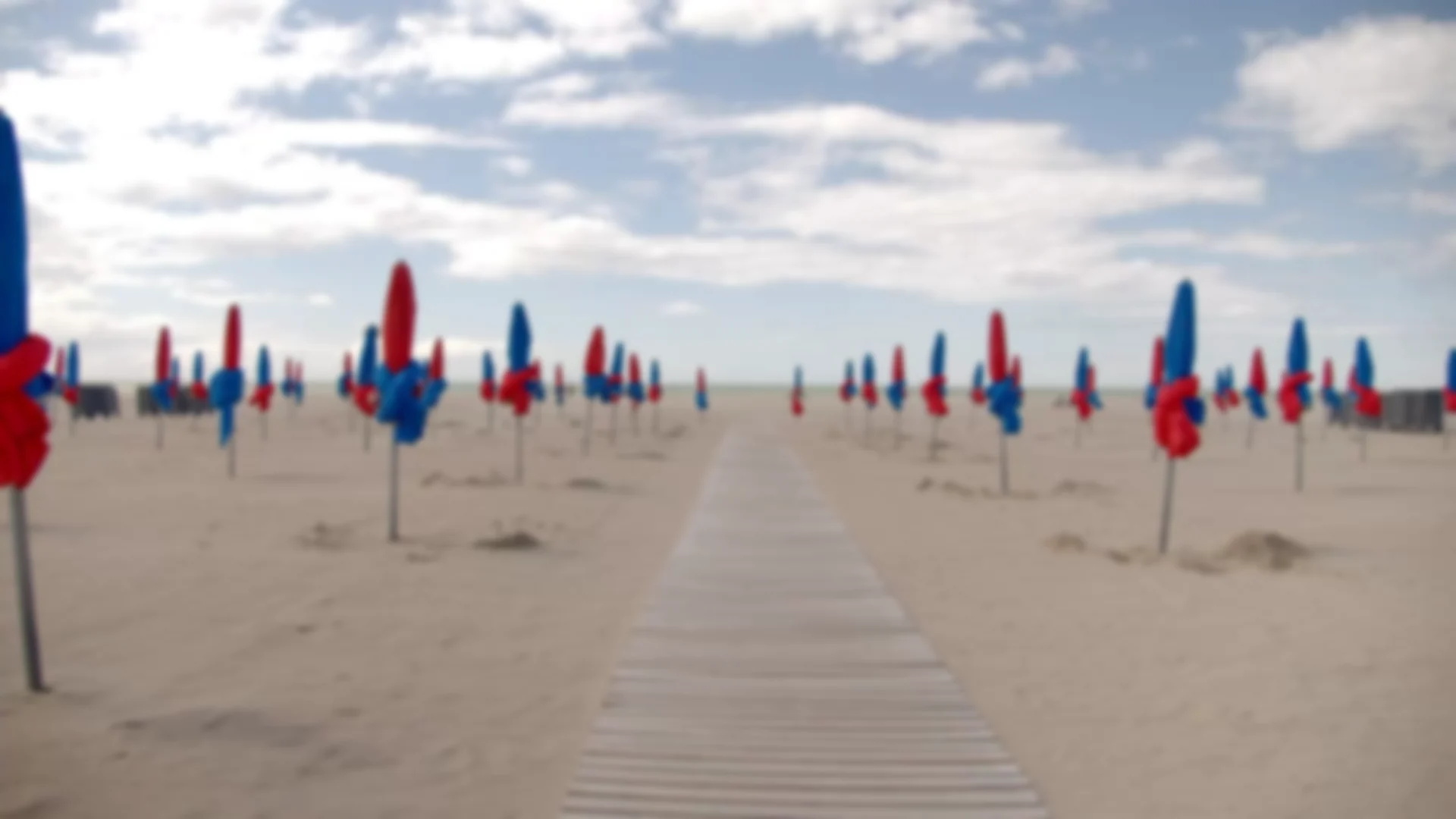 La plage de Deauville et ses parasols rouges et bleus repliés, plantés de part et d'autre d'un caillebotis de bois qui file droit vers la mer.