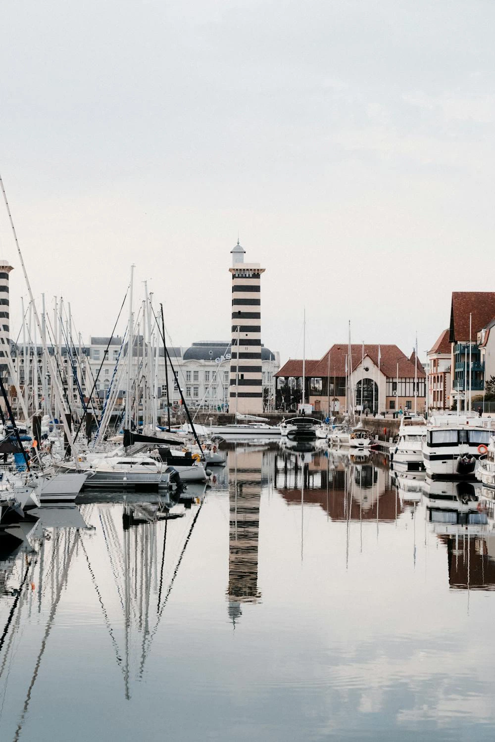 Le port de plaisance de Deauville sous un ciel bas : voiliers à quai, phare rayé noir et blanc et façades normandes reflétés dans l'eau immobile.
