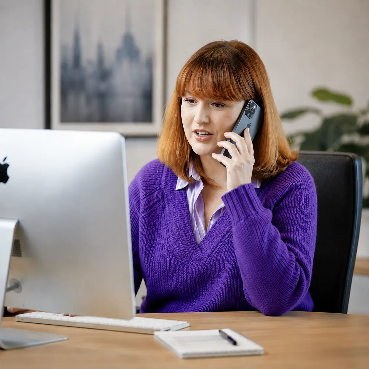 Une collaboratrice au téléphone à son bureau, devant un écran d'ordinateur, un carnet et un stylo posés à côté du clavier.