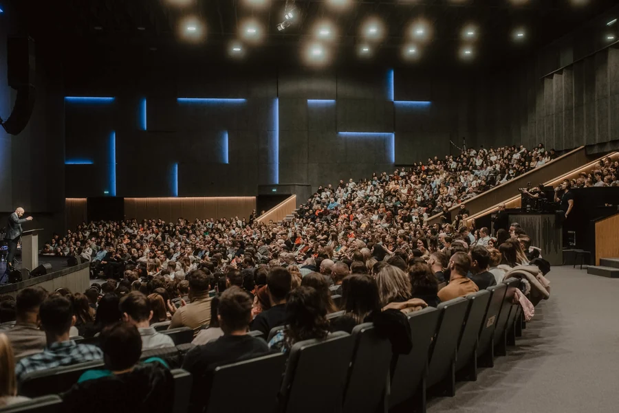 Plénière de convention dans un amphithéâtre comble : gradins occupés jusqu'au fond, intervenant au pupitre et bandes lumineuses bleues sur les murs.