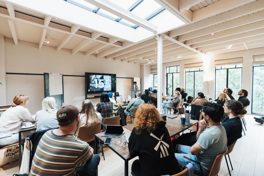 Journée d'étude dans une salle claire sous verrière : participants installés autour de tables basses, écran mural et intervenant debout devant l'assistance.