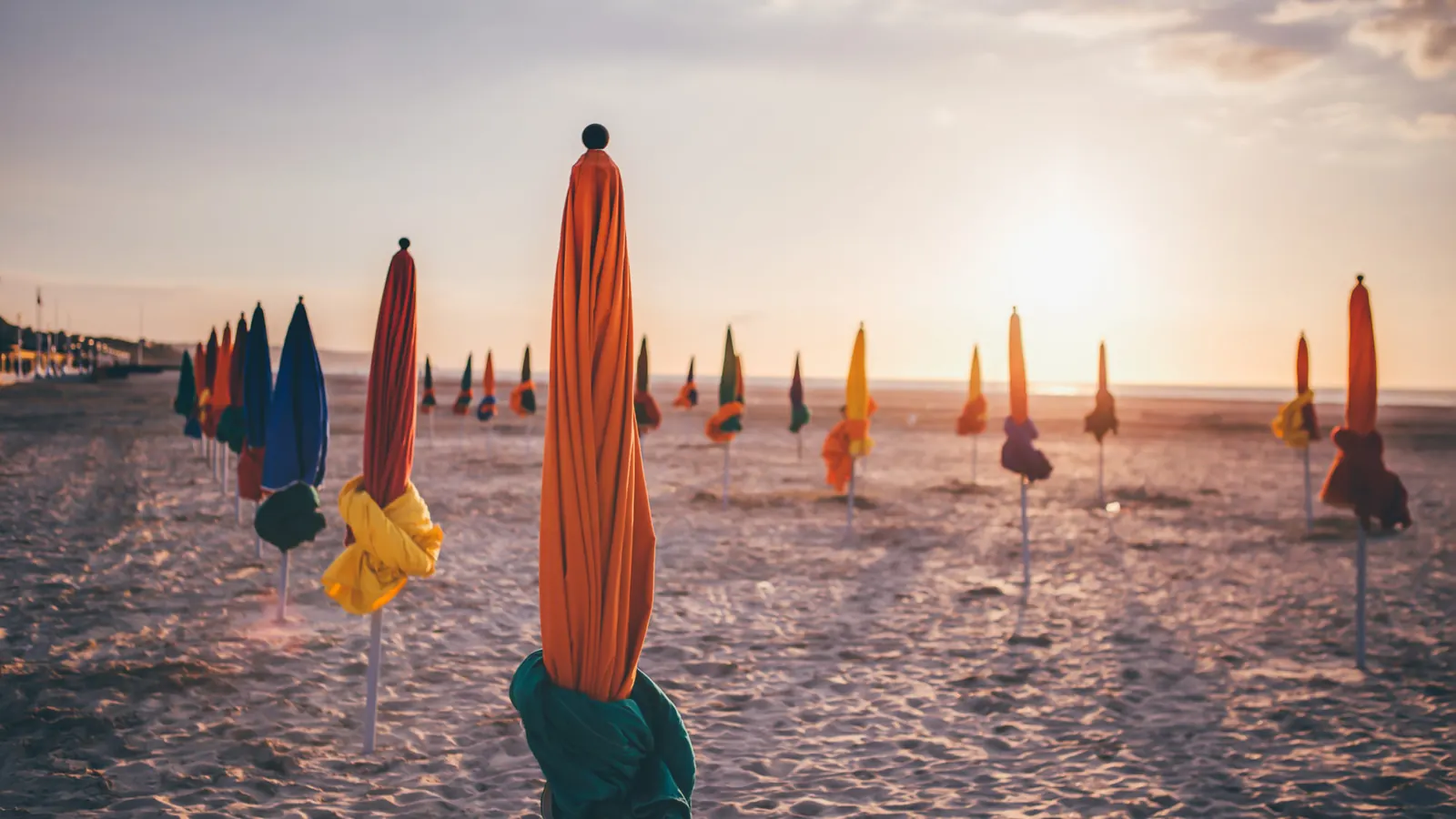 Parasols de Deauville fermés sur le sable, à contre-jour au soleil couchant ; un parasol orange se détache au premier plan, la mer et l'horizon en fond.