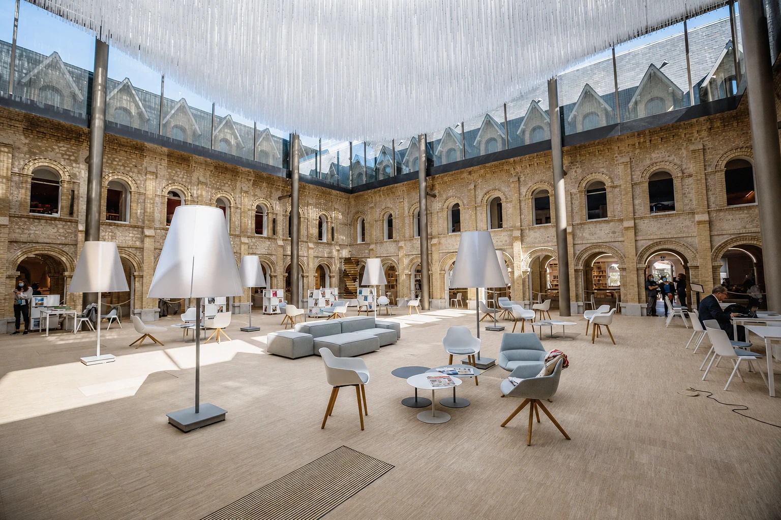 Le cloître vitré d'un ancien couvent reconverti : arcades de pierre claire sur deux niveaux, verrière et voile de fils suspendu, lampadaires blancs surdimensionnés et fauteuils bas répartis sur le sol de bois.