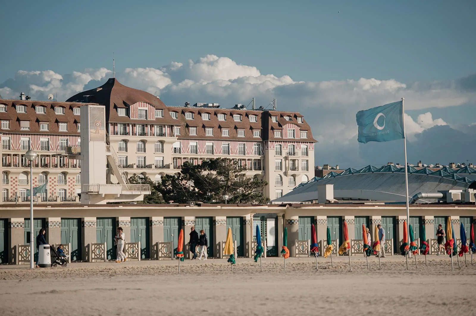 Un grand hôtel de front de mer aux façades à colombages rouges et blancs, vu depuis la plage, derrière l'alignement des cabines de bain et les parasols repliés des Planches.