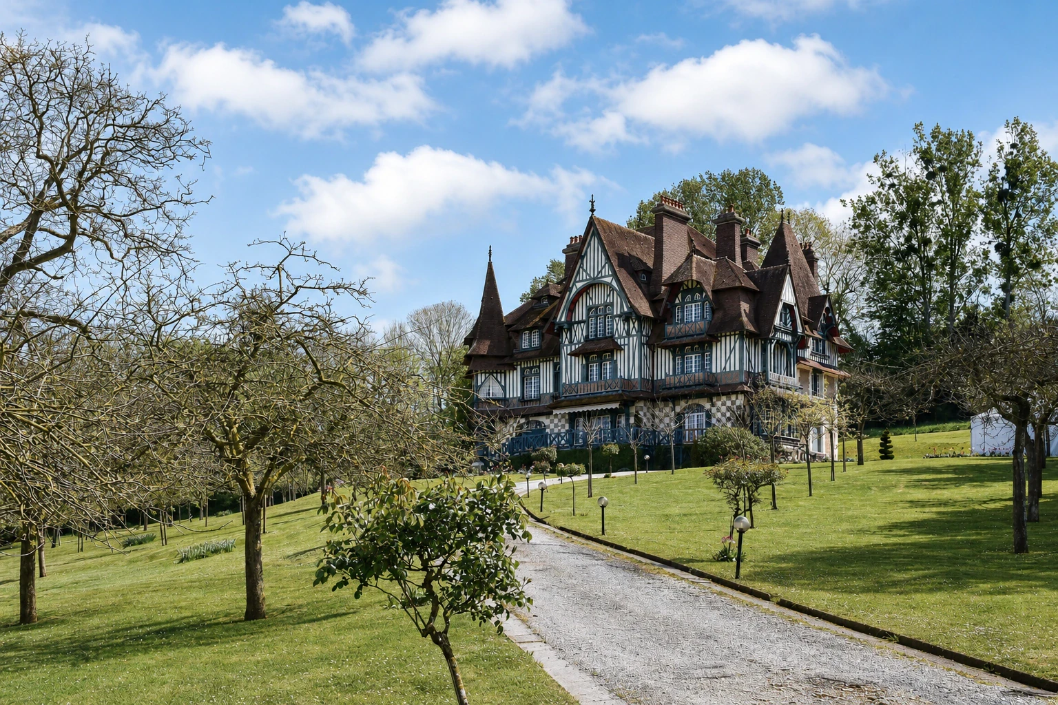 Un manoir normand à colombages et tourelles au bout d'une allée de gravier, entouré d'un parc arboré, sous un ciel de printemps.