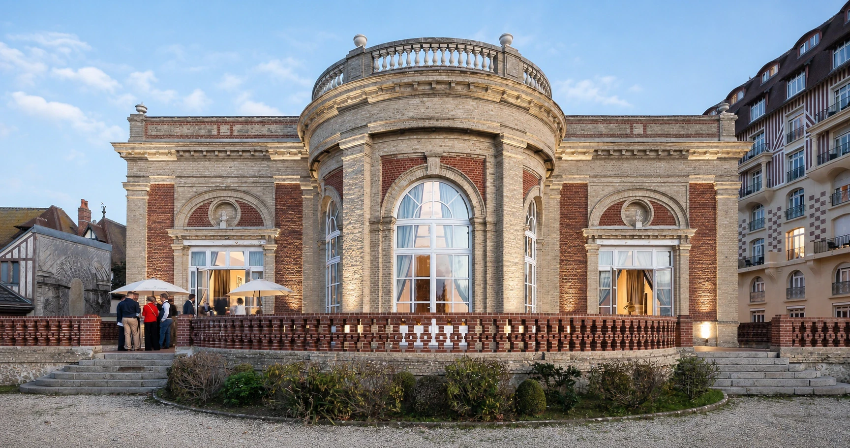 Une villa balnéaire en brique et pierre, rotonde centrale et hautes portes-fenêtres éclairées à la tombée du jour, quelques invités sur la terrasse.