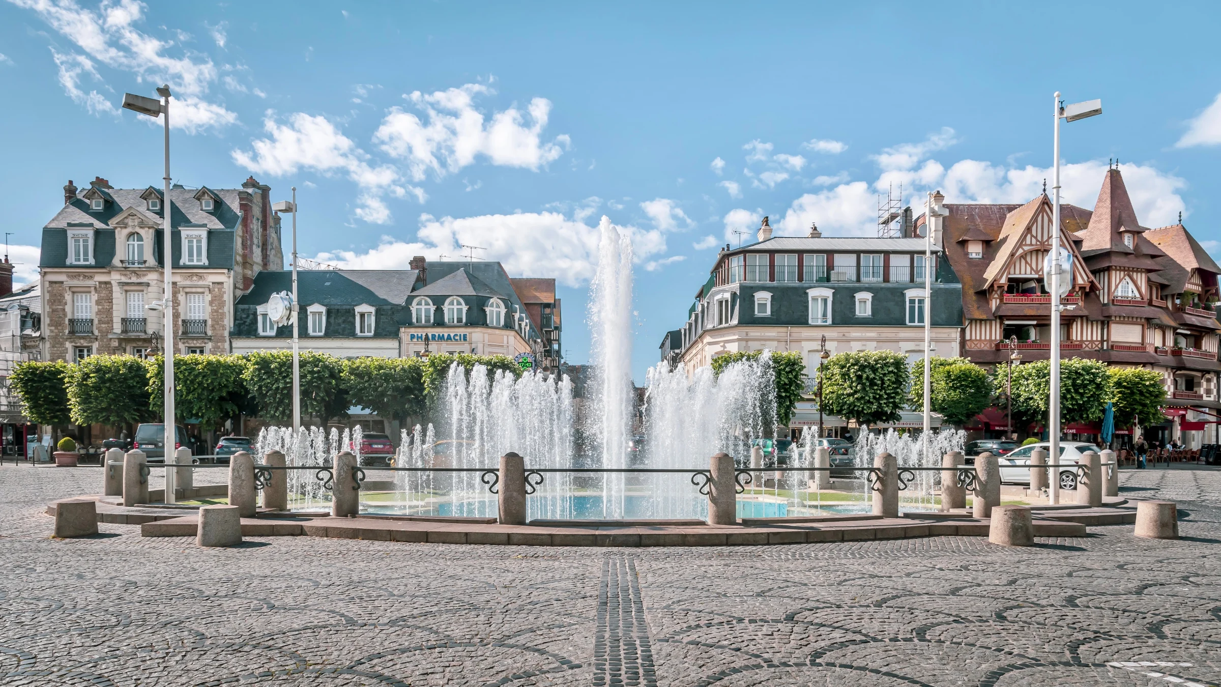 La fontaine de la place Morny en fonctionnement, au centre de Deauville, entourée de son pavage en éventail et des façades à colombages et toits d'ardoise.