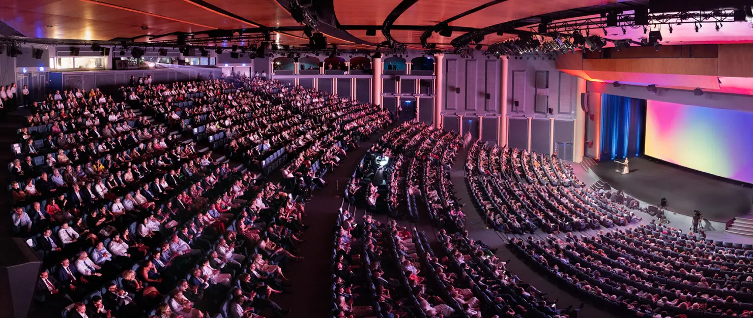 Une grande salle de spectacle de Deauville pendant une séance : gradins occupés sur plusieurs niveaux, écran géant éclairé et scène au fond.