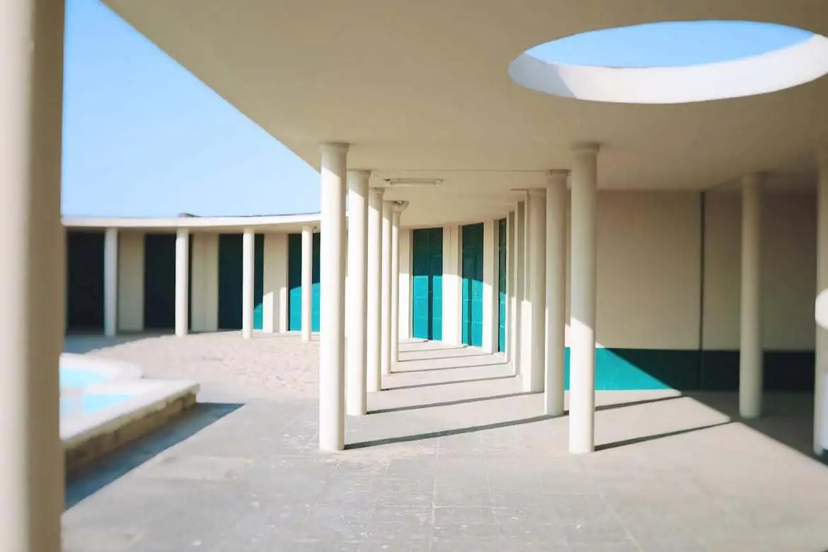 La colonnade blanche d'un établissement de bains de Deauville : cabines aux portes turquoise, sol de sable clair et oculus percé dans le plafond.