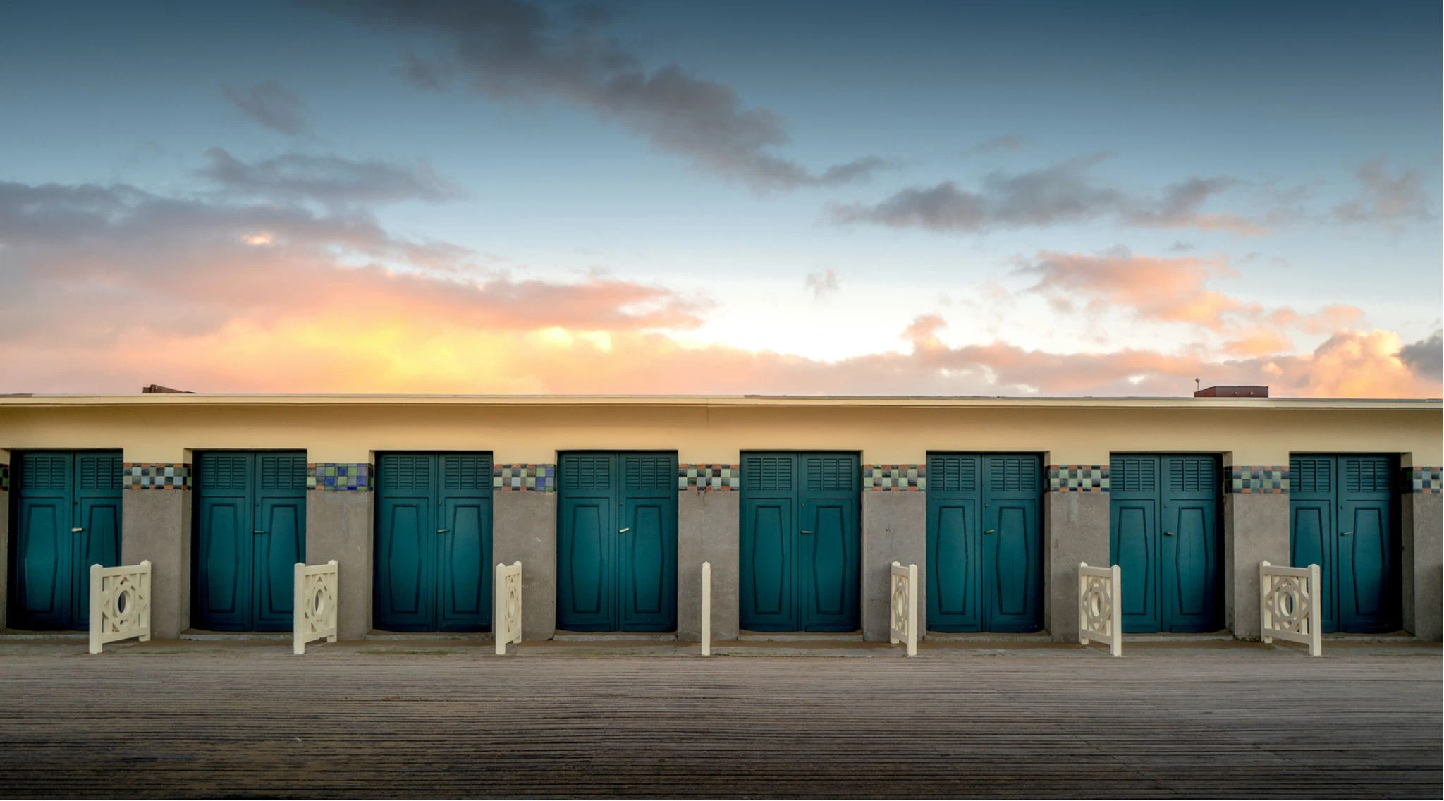 La rangée de cabines des Planches de Deauville au coucher du soleil : portes de bois turquoise, frise de céramique colorée et promenade de planches au premier plan.