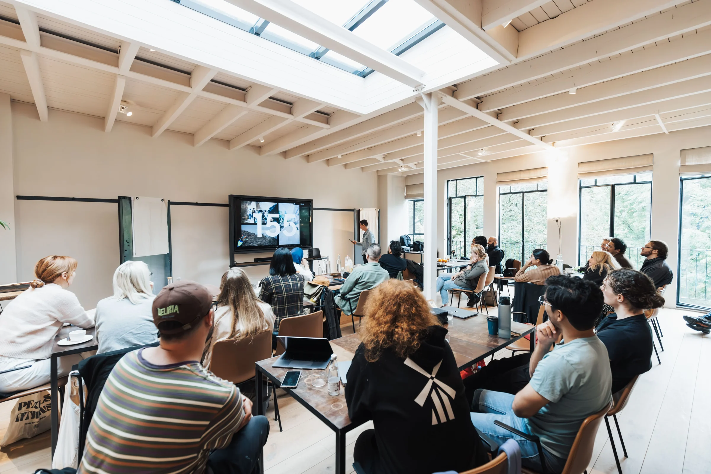 Journée d'étude dans une salle claire sous verrière : participants installés autour de tables basses, écran mural et intervenant debout devant l'assistance.
