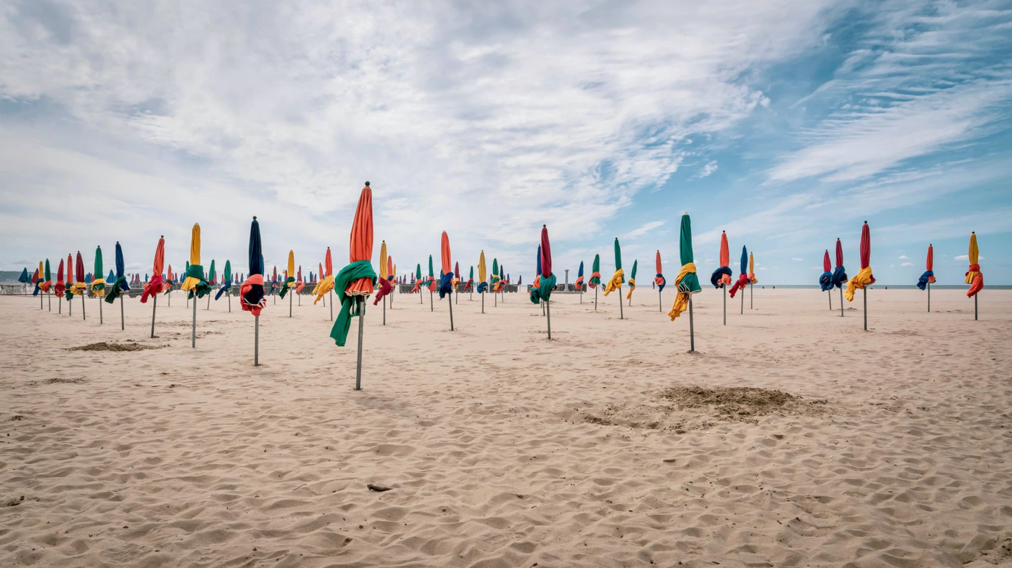 Des dizaines de parasols fermés, rouges, verts, bleus et jaunes, plantés dans le sable de la plage de Deauville sous un ciel nuageux.
