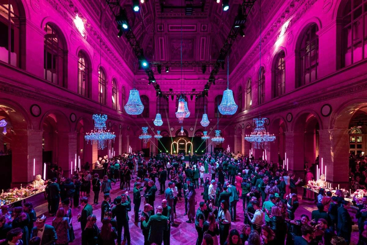 Une soirée de gala dans une grande salle sous éclairage violet : lustres de cristal suspendus, plusieurs centaines d'invités en tenue de soirée et une scène éclairée au fond.