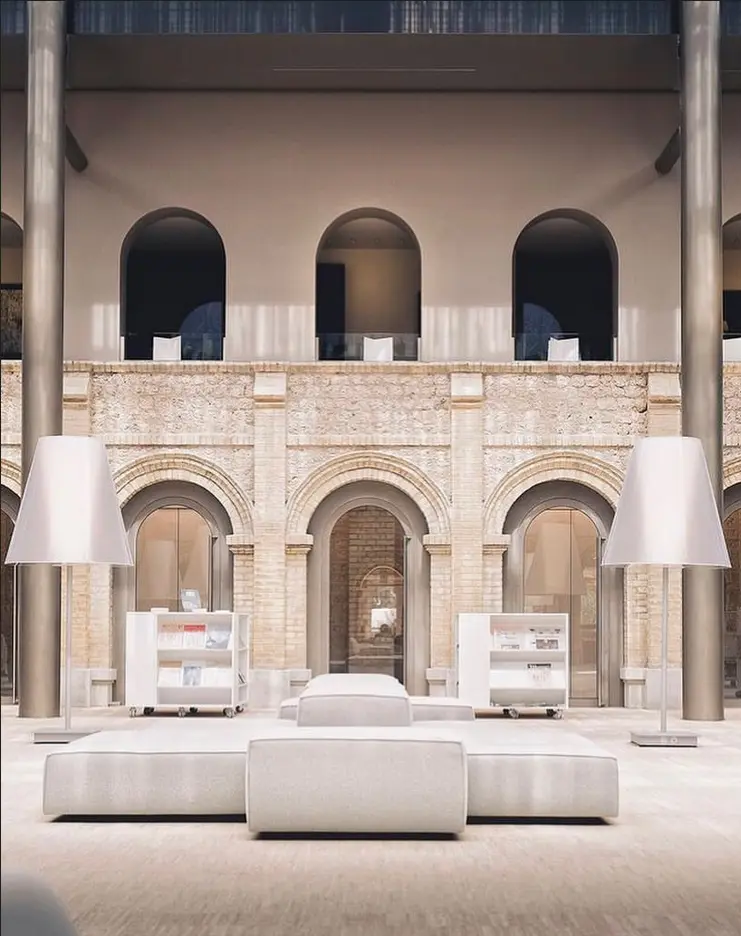 Arcades de pierre claire d'un cloître reconverti en espace de réception : galerie à l'étage, très grandes liseuses blanches et banquettes basses au centre.