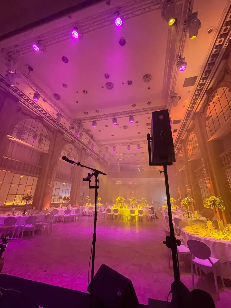 Vue depuis la scène d'un dîner de gala : tables rondes nappées de blanc et hauts bouquets, sous des projecteurs magenta et ambrés accrochés à une structure, dans une salle à hautes fenêtres cintrées.