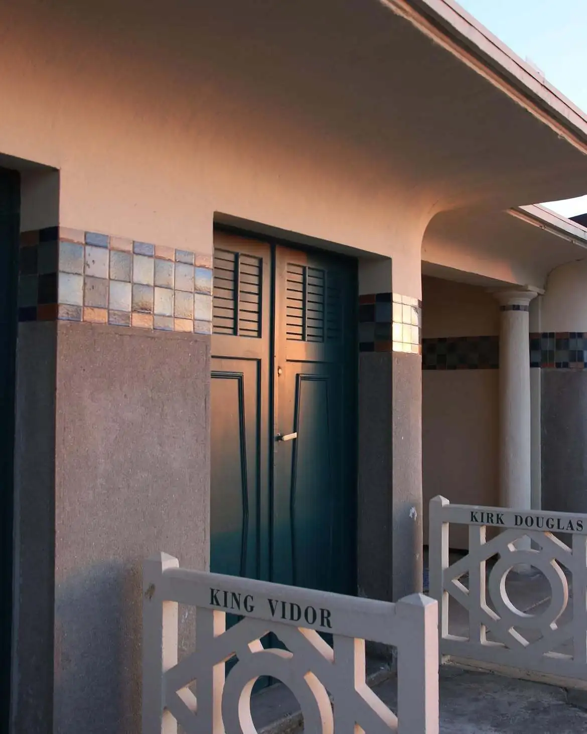 Une cabine des Planches de Deauville en fin de journée : porte turquoise à persiennes et barrières blanches portant les noms de King Vidor et Kirk Douglas.
